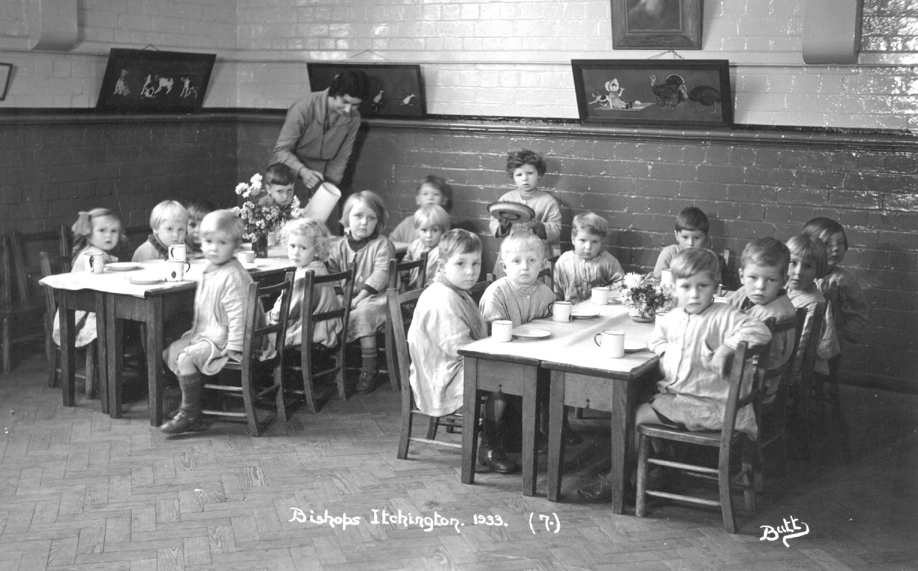Black and white image of children sat at tables, all looking at the camera. Taken 1933