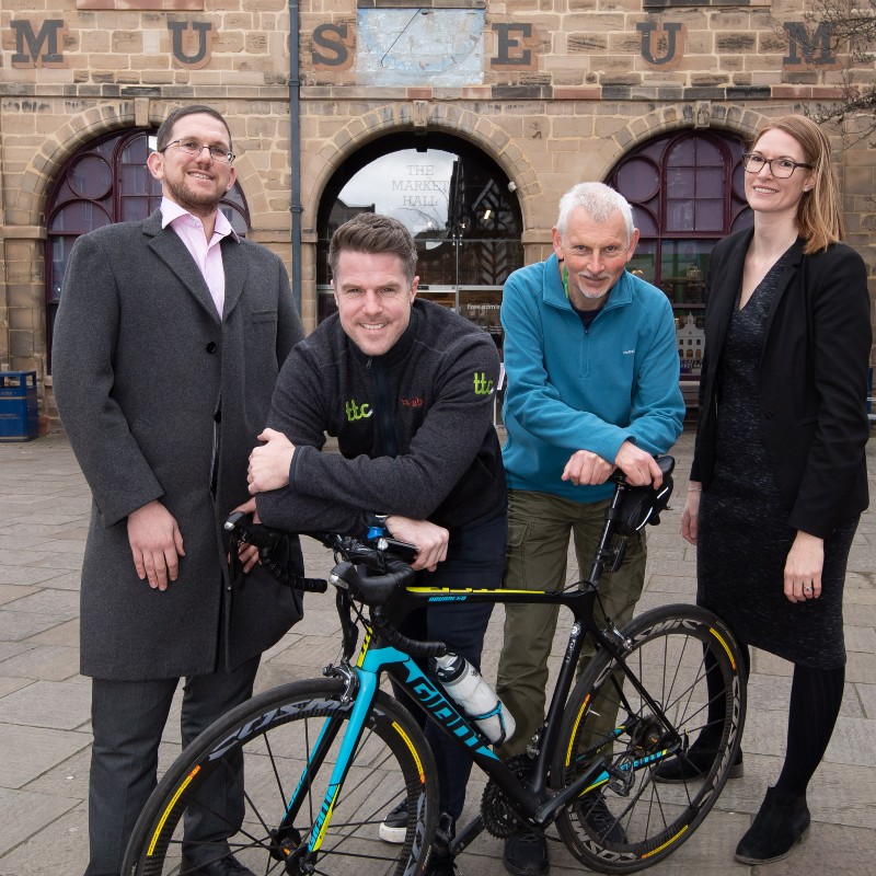 Two men in sweaters leaning on a bike with a man and woman stood either side in suits. All stood in front of the Warwickshire museum.