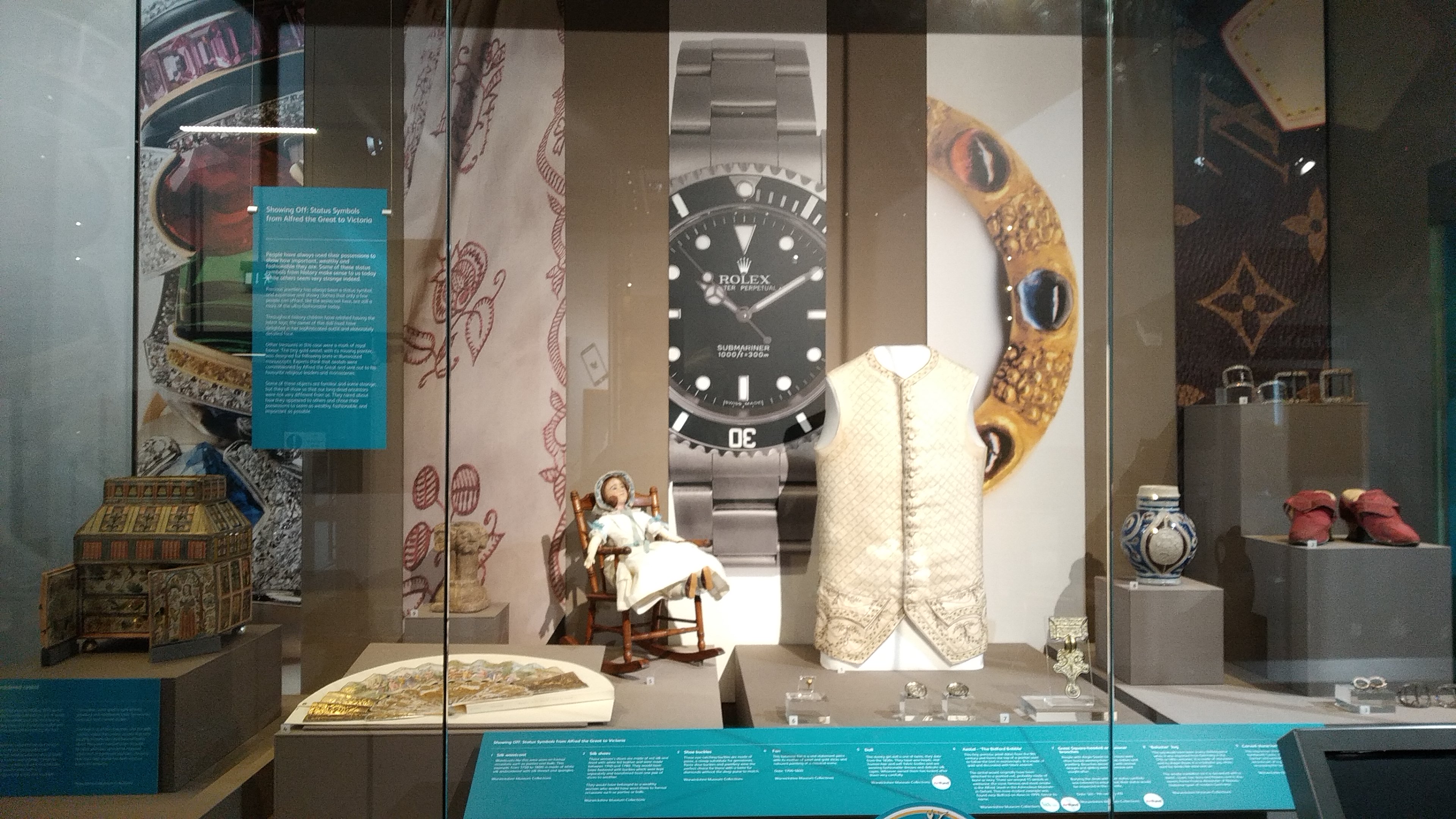 photograph of new display at market hall museum. the display is in a glass case holidng a golden button vest, a vase and a doll among other artefacts.