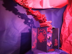 A photo of an intricate red design on a black box which has red rolled tissue paper coming from the top of the box. 